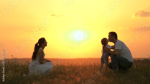 little daughter goes from mom to dad, hugs and kisses her parents in rays of warm sun. Happy family walks in park at sunset. Mom, dad and baby. healthy family plays in field. Happy family concept