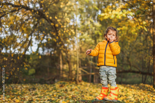 Little funny girl in stylish yellow raincoat and rubber boots stands in the autumn forest or park outdoors. Back profile side-view. Maple leaves foliage on the ground. Concept fall family photo shoot
