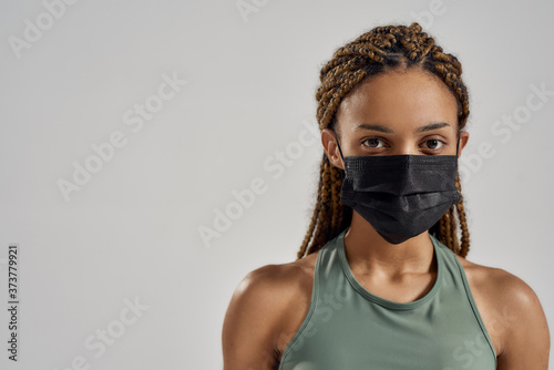 Portrait of a young sportive mixed race woman wearing black medical protective face mask looking at camera while standing isolated over grey background photo