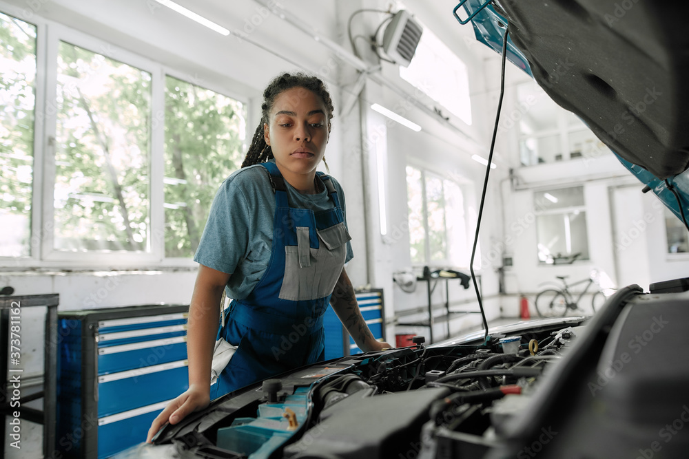 For better ride. Young african american woman, professional female mechanic looking, examining under hood of car at auto repair shop For better ride. Young african american woman, professional female mechanic looking, examining under hood of car at auto repair shop