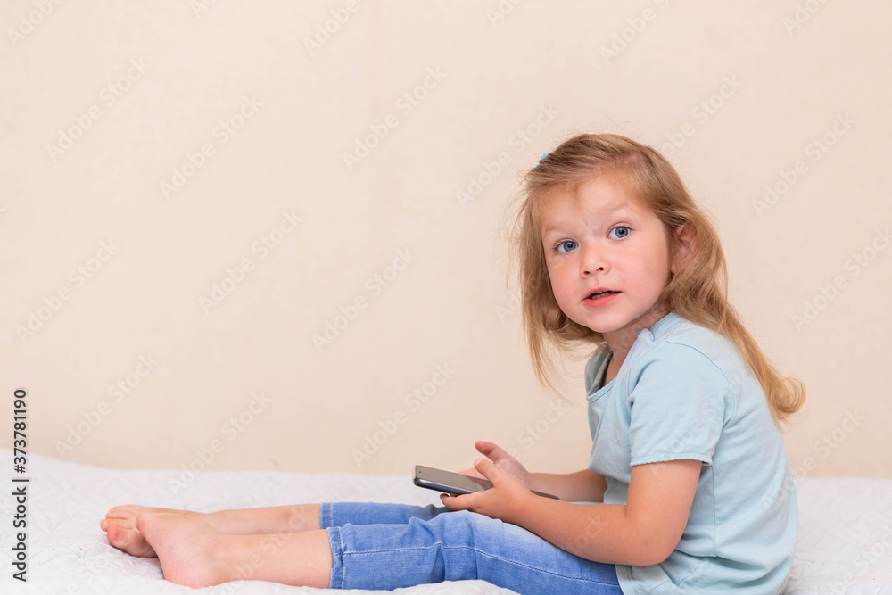 Curious little girl in blue clothes, uses phone applications,looking at a mobile device, sits on a bed.Clear girl, holds a smartphone, plays a game online alone at home.Concept of a child and a gadget