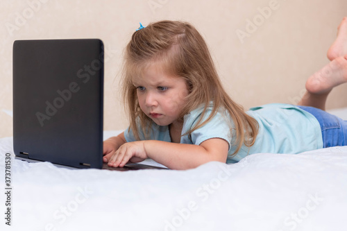 Little girl in blue clothes lies on the bed and types, plays on a laptop computer. Concept for children's internet safety, social networks or education and homework