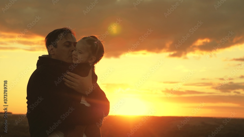 Happy child and father are playing in field in sun. dad hugs and covers his little daughter with jacket on cool evening in park at sunset. healthy baby kisses daddy. happy family and childhood concept