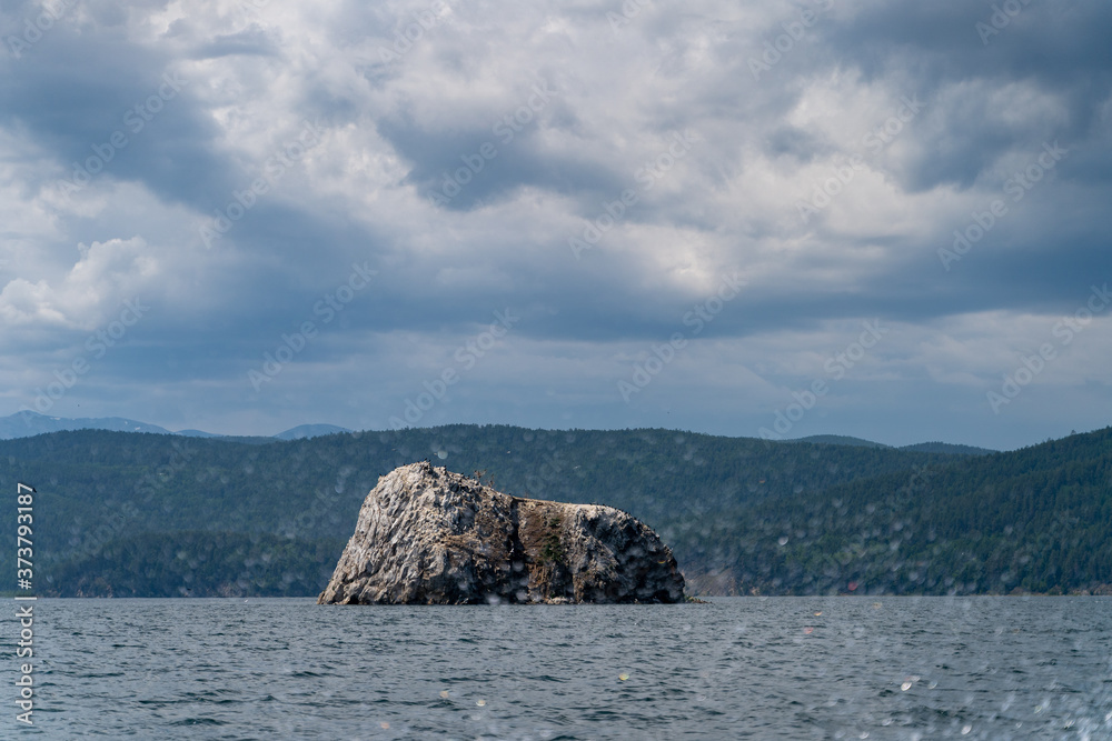 Russia, Irkutsk region, Baikal lake, July 2020: giant grey rock in the centre of lake with dramatic clouds above