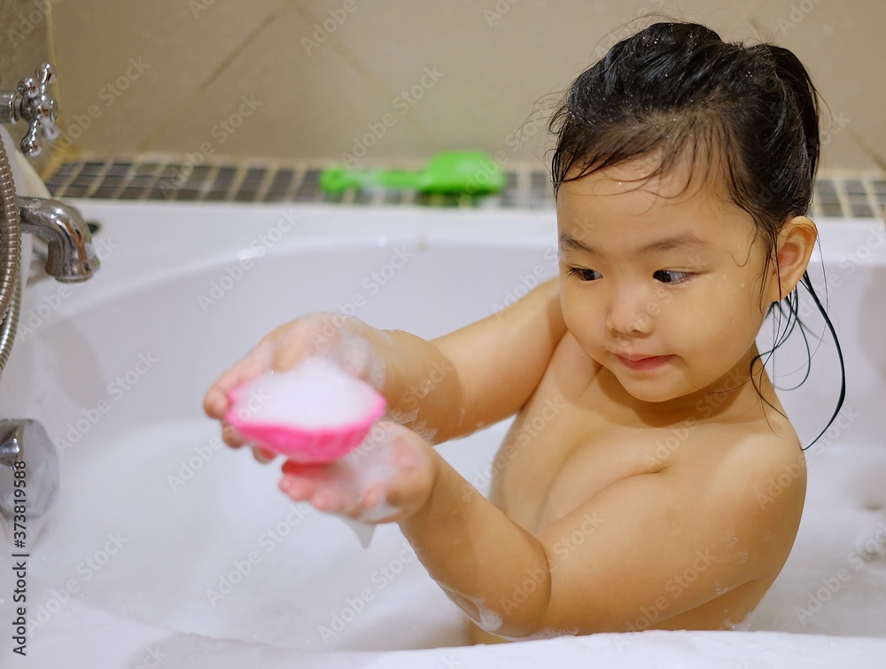 A cute young Asian girl playing with her pink plastic toy while taking a bubble bath in a white bathtub with foam and water. A cute young Asian girl playing with her pink plastic toy while taking a bubble bath in a white bathtub with foam and water.