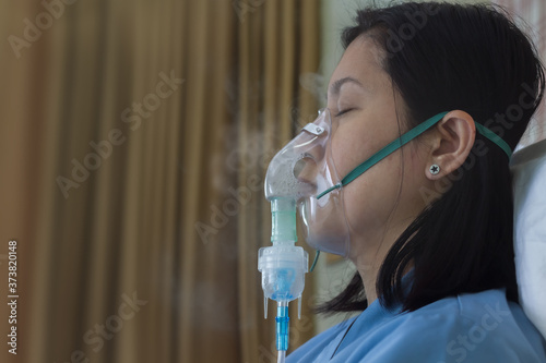 Sickness female in blue clothe wearing nasal mask with respiratory problem in hospital room. Asian woman patient inhalation therapy by the mask of inhaler with soft stream smoke from bronchodilator.