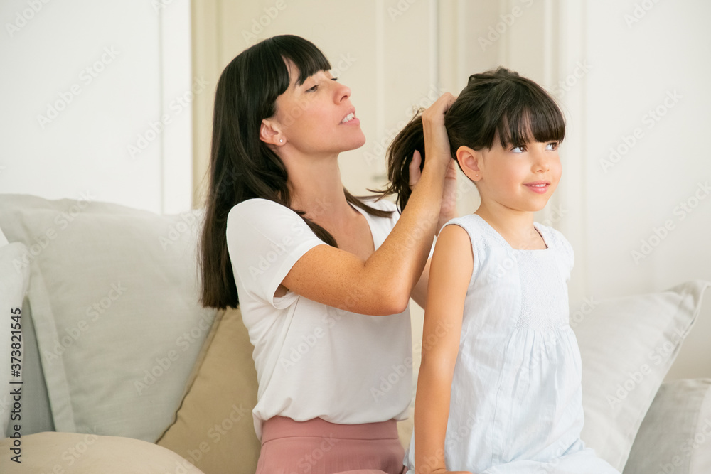 Young Caucasian mother holding hairs of her daughter. Pretty brunette mom sitting on sofa and doing ponytail hairstyle for little cute girl. Family time and parenthood concept Young Caucasian mother holding hairs of her daughter. Pretty brunette mom sitting on sofa and doing ponytail hairstyle for little cute girl. Family time and parenthood concept