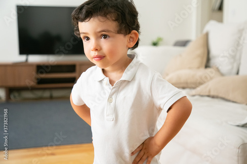 Adorable little boy in white shirt standing with hands on hips in living room. Lovely child posing and looking away. Blurred background. Portrait and appearance concept