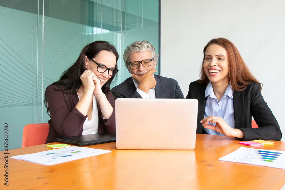 Happy business team watching successful project presentation on laptop or having video call, sitting at meeting table with paper reports and charts. Medium shot. Communication concept Happy business team watching successful project presentation on laptop or having video call, sitting at meeting table with paper reports and charts. Medium shot. Communication concept