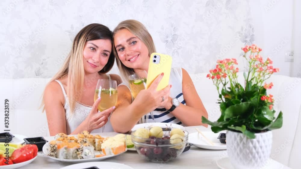  friendship and party concept - two young beautiful girlfriends drink wine and take selfie on smartphone in a cozy kitchen at the table, selective focus