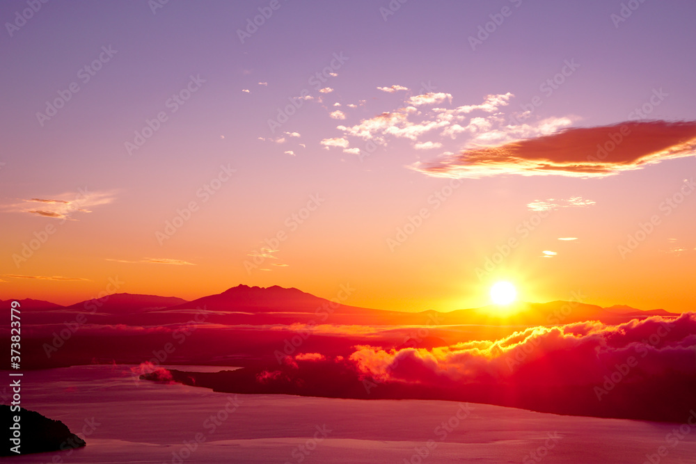 Obraz premium 夜明けの空に力強く輝く太陽。津別峠、北海道。Abstract of sunrise and sunset. The sun is shining in the sky,clouds flowing low,lake surface seen in thin light.Scenic view from high place. Tsubetsu-pass,hokkaido,japan.