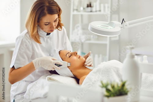 Young cosmetologist or dermatologist making ultrasound facial cleaning for woman in beauty salon photo