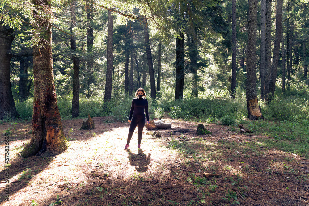 A female in a mask, a sports jacket alone is standing in a pine forest A female in a mask, a sports jacket alone is standing in a pine forest