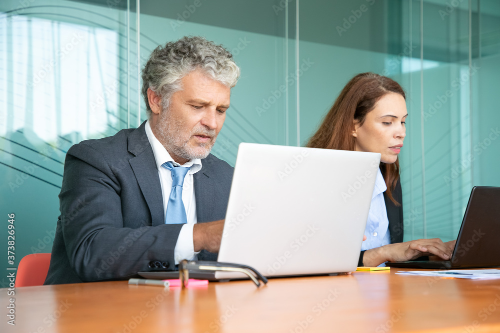 Two colleagues sitting together and using computers in office. Employees of different ages typing on laptop keyboards. Medium shot. Communication or wireless technology concept Two colleagues sitting together and using computers in office. Employees of different ages typing on laptop keyboards. Medium shot. Communication or wireless technology concept