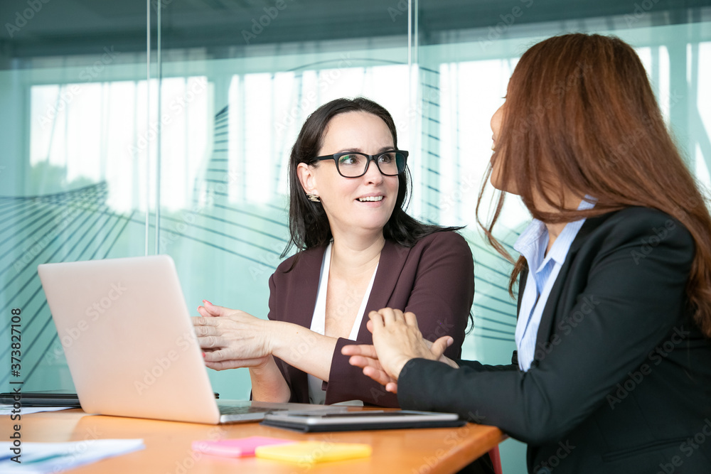 Excited cheerful businesswomen discussing project while sitting at open laptop, talking and smiling. Business communication and teamwork concept Excited cheerful businesswomen discussing project while sitting at open laptop, talking and smiling. Business communication and teamwork concept