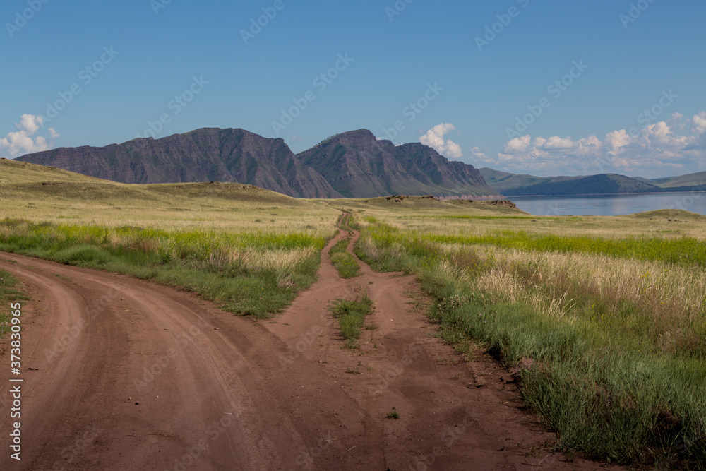 Summer landscape with a road among thickets of grass, a river and mountains in the background. The beauty of the surrounding world. Summer landscape with a road among thickets of grass, a river and mountains in the background. The beauty of the surrounding world.