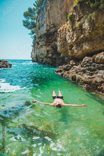 young man swimming on back in sea at bay between cliffs