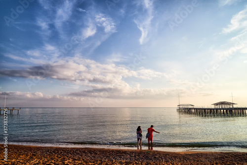 View from sand beach to water of sea, pier and silhouette of people or tourists in a nice day or evening with blues sky and white clouds.