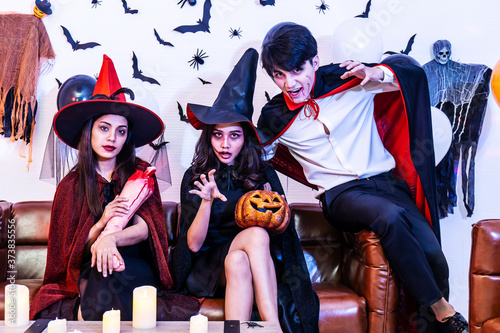 Asian Happy friends sitting on sofa in costumes and makeup on a celebration of Halloween posing with pumpkin and fake human leg in party, Group of young people enjoying Halloween party at home 
