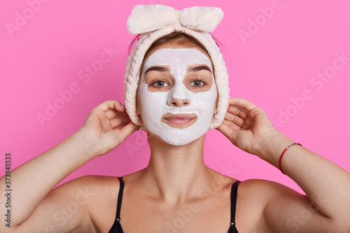 Attractive woman posing isolated over pink studio background, looking directly at camera, keeping hands on headband, stands with moisturizing mask.