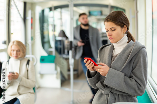 Young woman wearing light overcoat absorbed in her smartphone during trip in public transport
