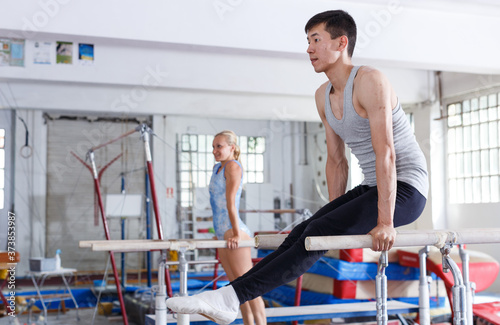 Man gymnast training gymnastic action at steel bars in gym, woman on background. High quality photo