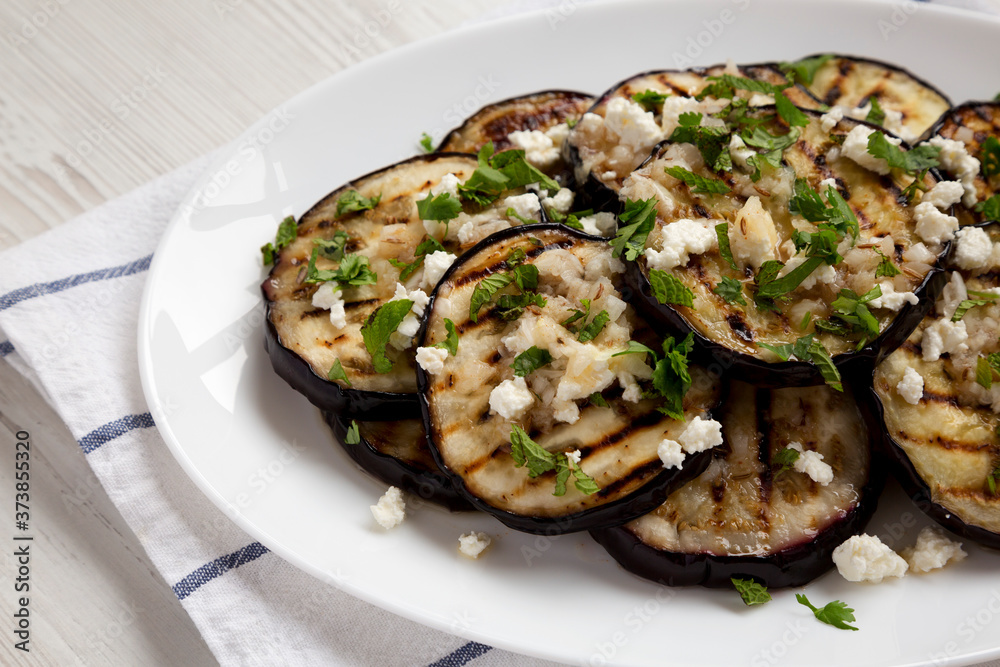 Homemade Grilled Eggplant with Feta and Herbs on a white plate on cloth, low angle view. Close-up.