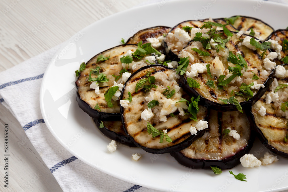 Homemade Grilled Eggplant with Feta and Herbs on a white plate on cloth, low angle view. Close-up. Homemade Grilled Eggplant with Feta and Herbs on a white plate on cloth, low angle view. Close-up.