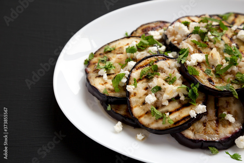 Homemade Grilled Eggplant with Feta and Herbs on a white plate on a black surface, side view. Copy space.
