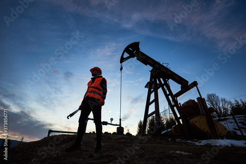 Horizontal snapshot of a silhouette of an oilman in orange vest and helmet, standing with his back to the oil pump jack in oilfield holding a pipe wrench, looking away, evening sky on background