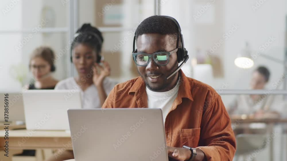 Cheerful Afro-American man in headset using laptop and speaking on web call while working in call center
