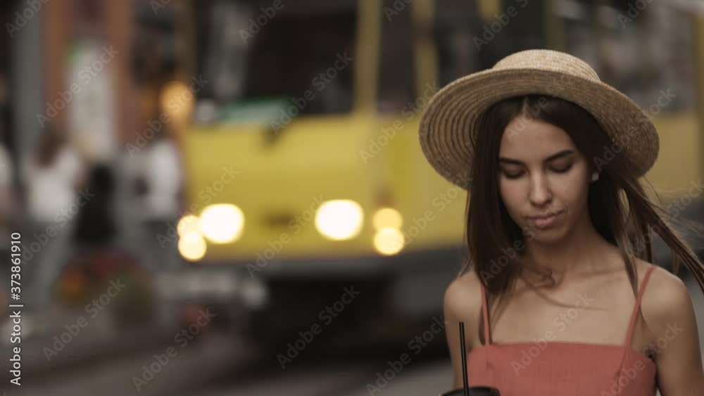 Beautiful young woman in a dress and hat walks around the city and enjoys a warm summer evening. An attractive girl drinks coffee and uses the phone while walking Beautiful young woman in a dress and hat walks around the city and enjoys a warm summer evening. An attractive girl drinks coffee and uses the phone while walking