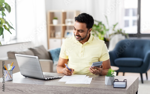 remote job, technology and people concept - young indian man with calculator and papers working at home office