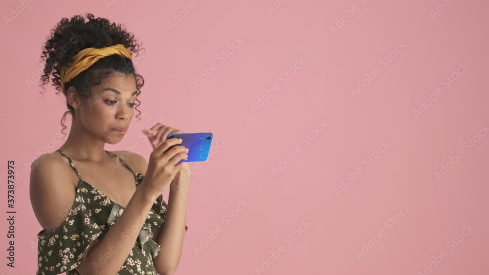 A beautiful african american young woman is watching video on her smartphone standing near free-space isolated over pink background A beautiful african american young woman is watching video on her smartphone standing near free-space isolated over pink background