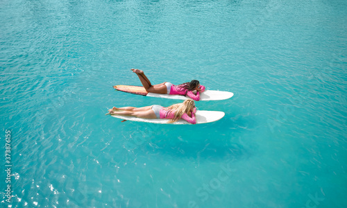 Blonde and brunette surfer girls on a long surfboards relax in the blue transparent ocean. From above aerial drone shot.
