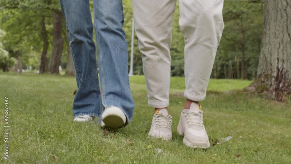 Close up of legs of unrecognizable man and woman wearing jeans and sneakers are walking on grass in park Close up of legs of unrecognizable man and woman wearing jeans and sneakers are walking on grass in park