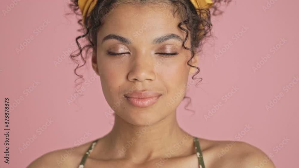 A close-up view of a happy african american young woman is opening her eyes and looking to the camera standing isolated over pink background A close-up view of a happy african american young woman is opening her eyes and looking to the camera standing isolated over pink background