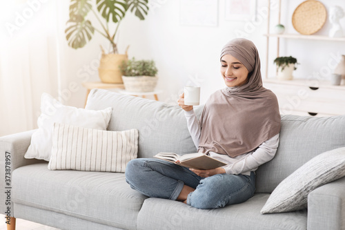 Happy Arabic Housewife Relaxing With Book And Coffee On Couch At Home