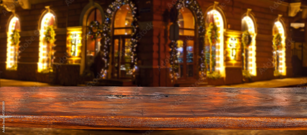 Old shabby board on a background of blurred Christmas market. Brown table top in sharpness. Christmas decorations, lights and lanterns in the background. Layout for design. Free space for products.