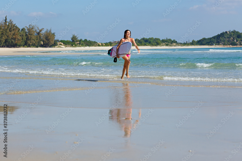 Fototapeta premium young slender and tanned girl in a swimsuit with a surfboard in her hands on the beach near the water, sea, summer, heat, sunny day, clear sea water, wave, lifestyle, sports, leisure, vacation, weeken