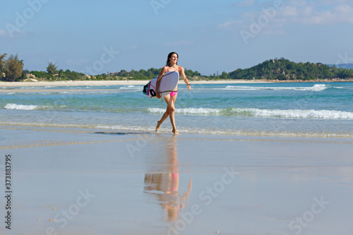 young slender and tanned girl in a swimsuit with a surfboard in her hands on the beach near the water, sea, summer, heat, sunny day, clear sea water, wave, lifestyle, sports, leisure, vacation, weeken
