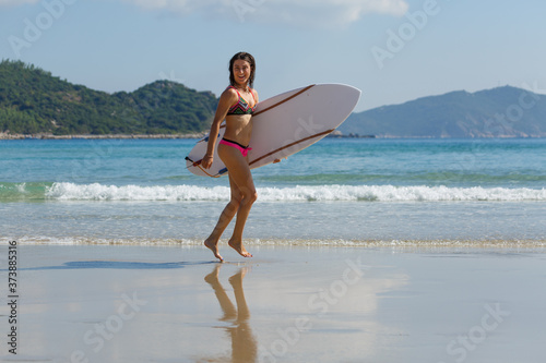young slender and tanned girl in a swimsuit with a surfboard in her hands on the beach near the water, sea, summer, heat, sunny day, clear sea water, wave, lifestyle, sports, leisure, vacation, weeken