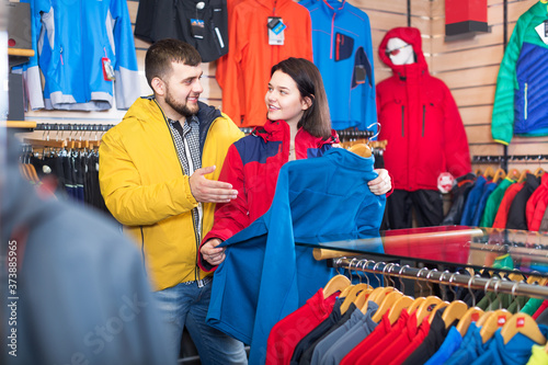 Young glad couple choosing track jacket in sports clothes store