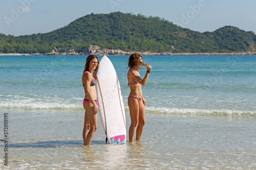 two girls in bekin with a white surfboard are standing near the sea, summer, heat, sunny day, clear sea water, wave