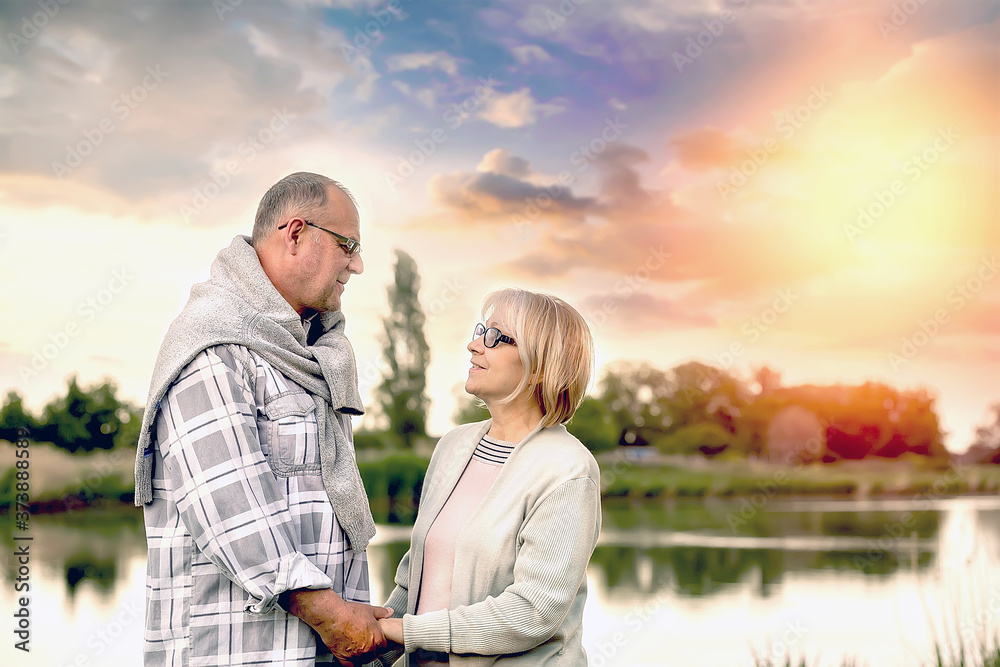A mature woman and a man in light-colored clothes look at each other with a smile and hold their hands in the form of a heart, People's life 60 plus. Sunset on the lake. Copy space on the left