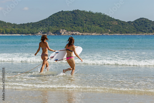 two girls in bekin with a white surfboard enter the blue sea, summer, heat, sunny day, clear sea water, wave