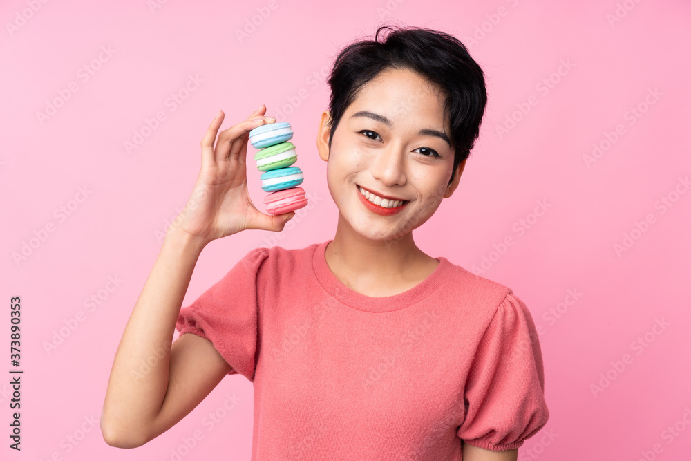 Young Asian girl over isolated pink background holding colorful French macarons and smiling a lot Young Asian girl over isolated pink background holding colorful French macarons and smiling a lot