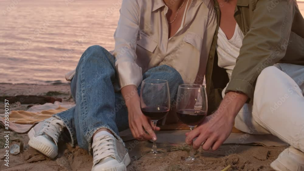 Medium shot of young loving spouses are sitting on plaid on sea coast at sunset, enjoying the view and drinking red wine