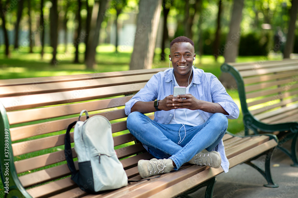 Fototapeta premium Cool black guy in earphones listening to music online from his smartphone on bench at park, empty space