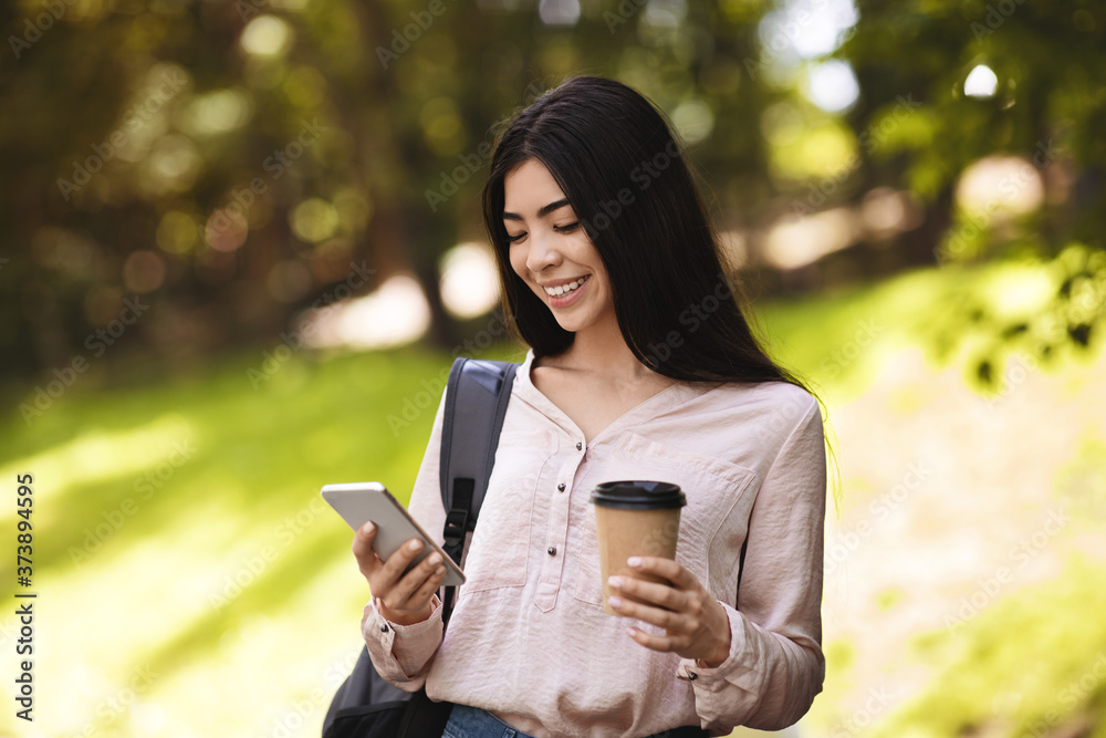 Asian Student Girl Using Smartphone While Walking With Backpack And Coffee Outdoors